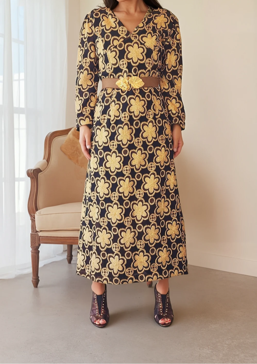 Model standing in a vintage 1970s midi dress featuring a bold black and gold floral pattern, styled with a statement belt and open-toe heels.