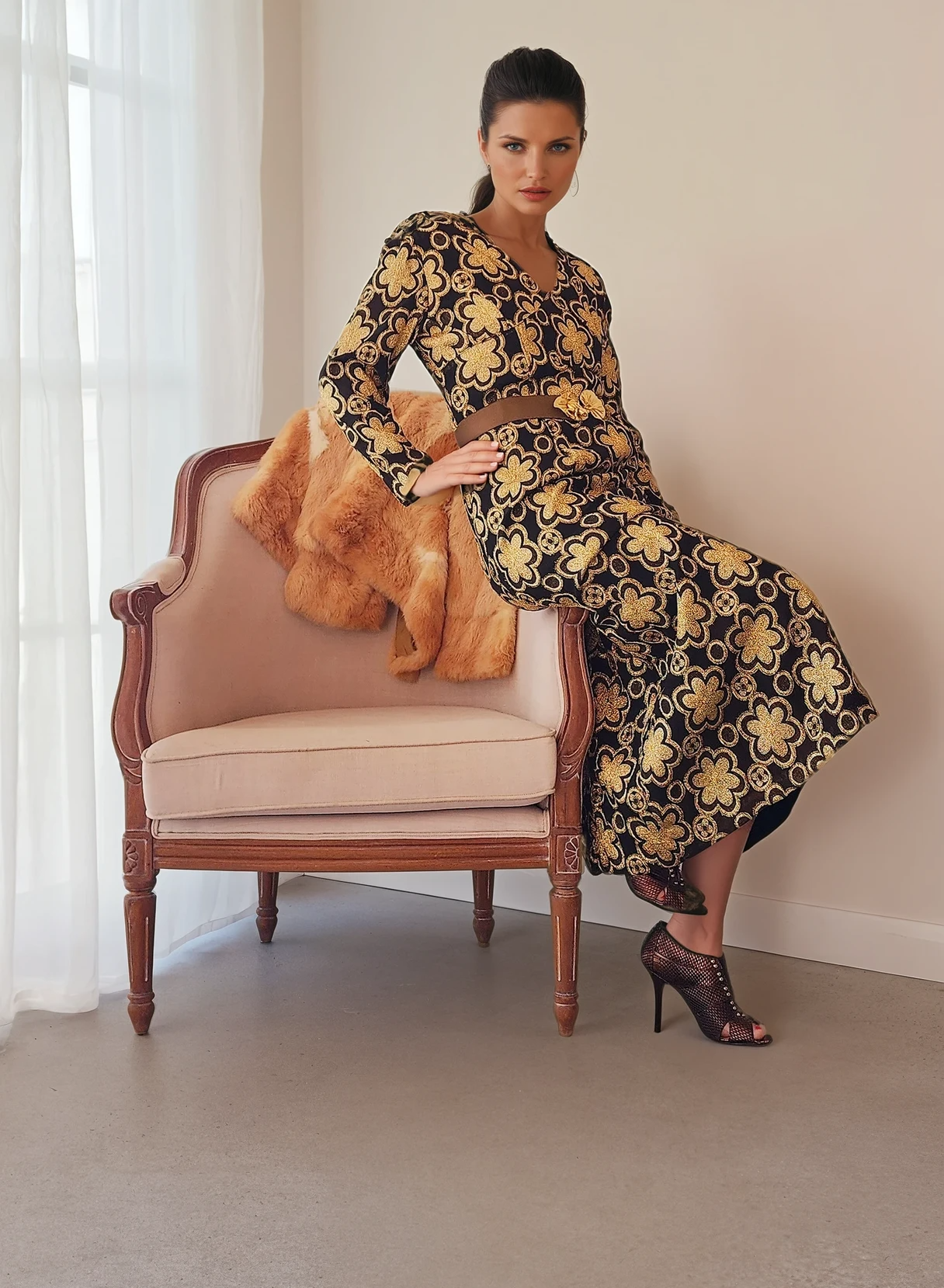 Model leaning against a velvet armchair, showcasing the vintage dress’s golden embroidery and rich texture in a relaxed pose