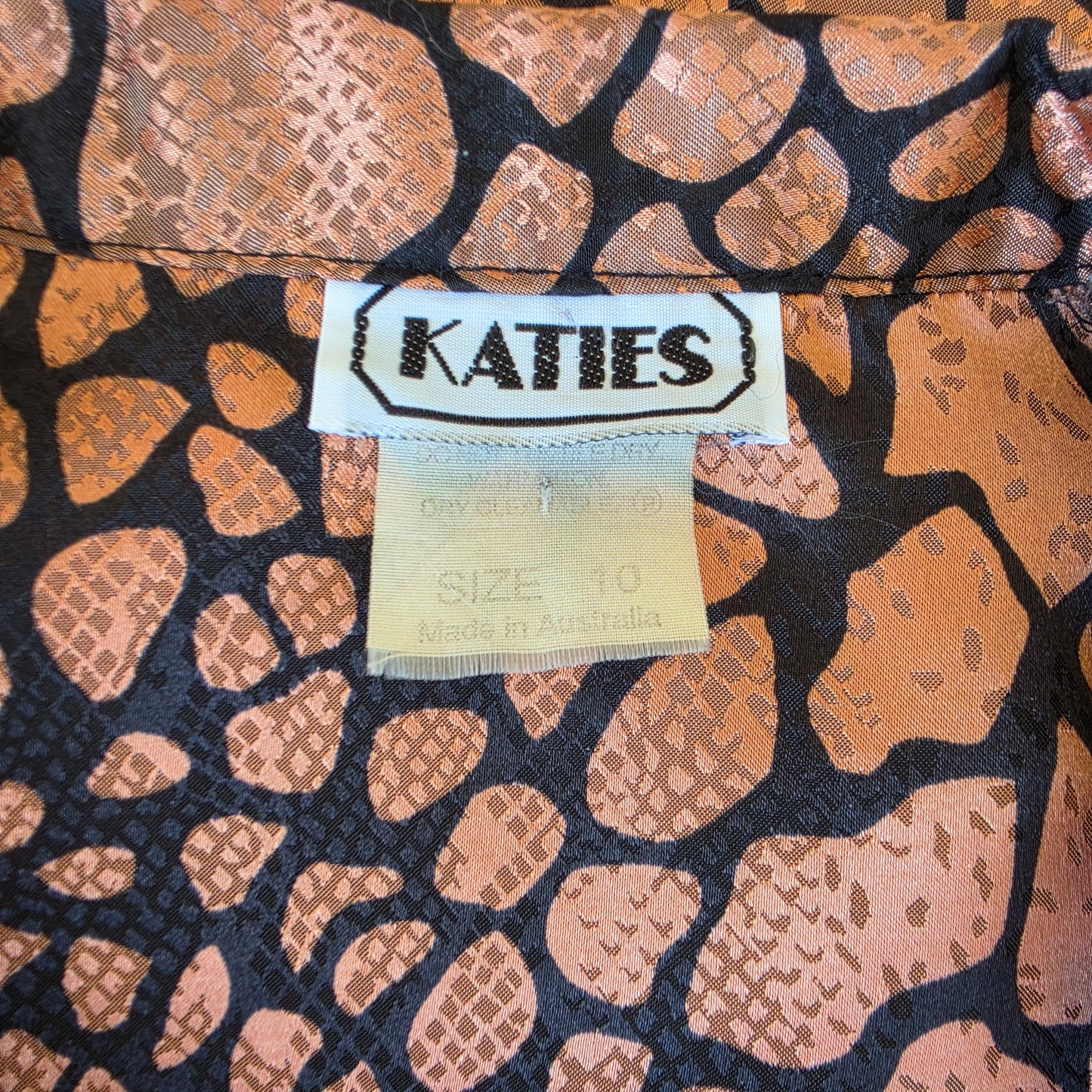 Close-up of a garment's inner neck label reading KATIES, showing a bronze and black animal-print fabric with a snakeskin pattern.