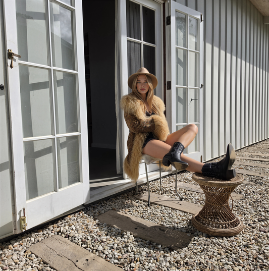 A woman sits on a metal chair outside a white building with French doors, wearing the vintage Tapestry Coat, a fedora hat, black shorts, and black ankle boots. A small woven stool sits beside her on a gravel surface.