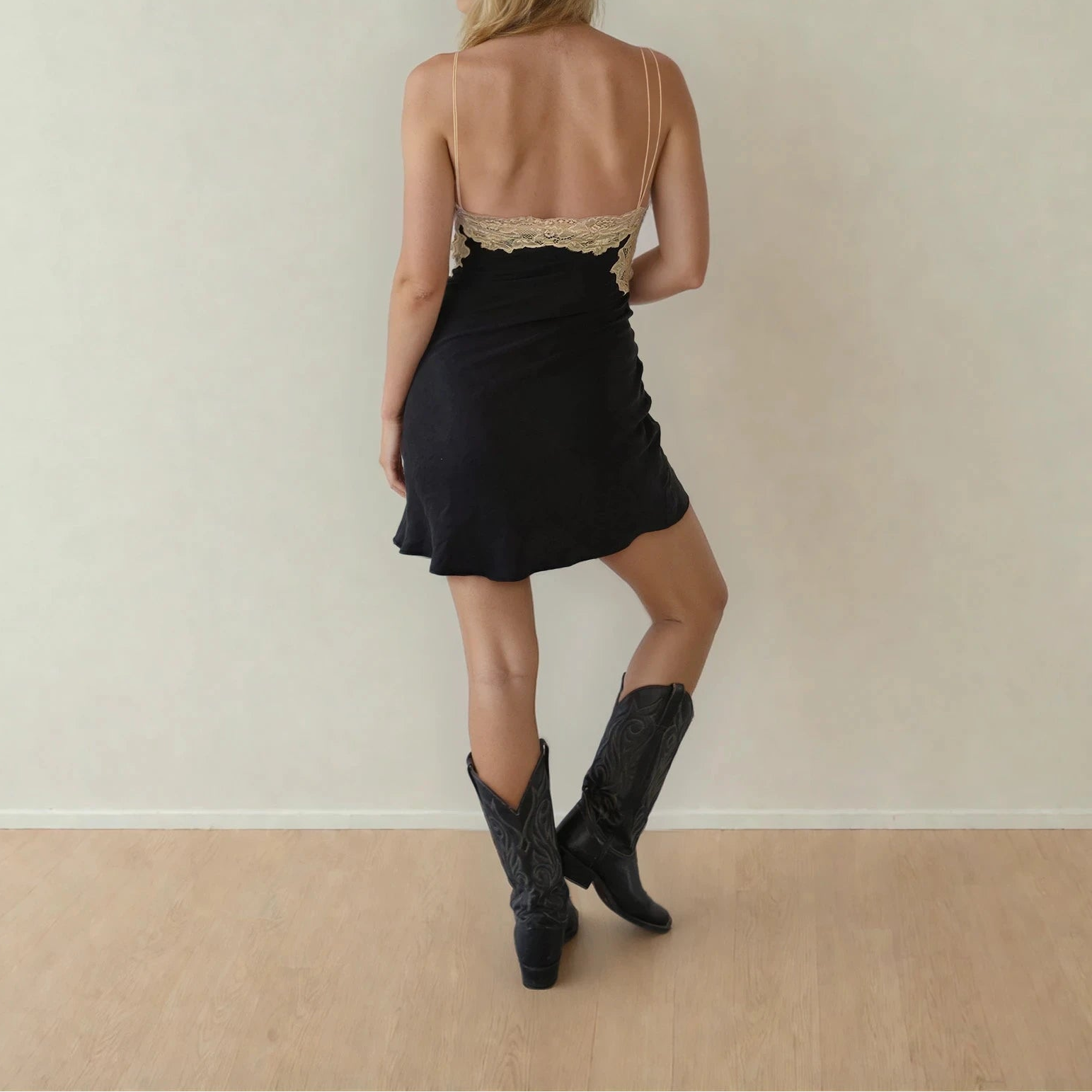 Woman wearing a black Verge Girl mini dress with lace detailing and black boots against a plain wall.