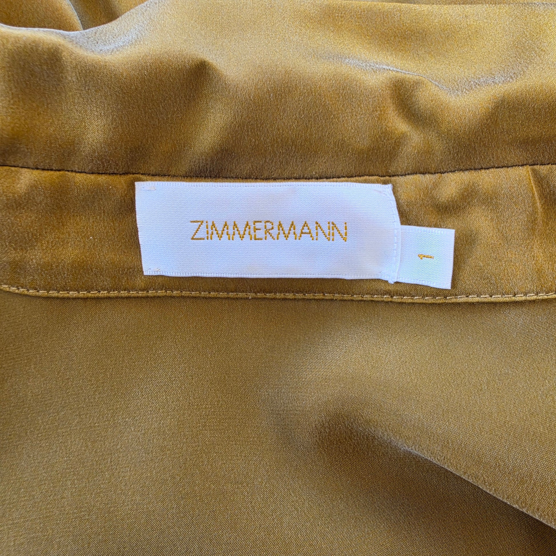Close-up of a white fabric label with the brand name ZIMMERMANN in gold lettering, sewn onto the gold silk wrap midi dress.