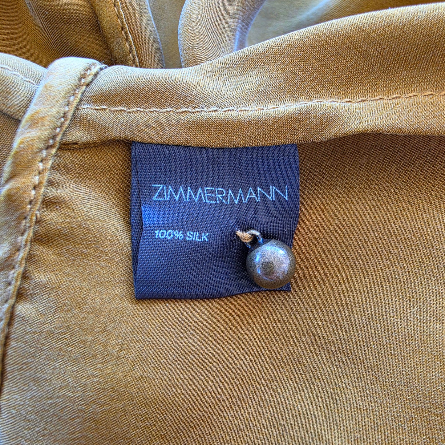 Close-up of the tan garment label reading ZIMMERMANN 100% SILK, with a small metallic pearl pin attached to the side.