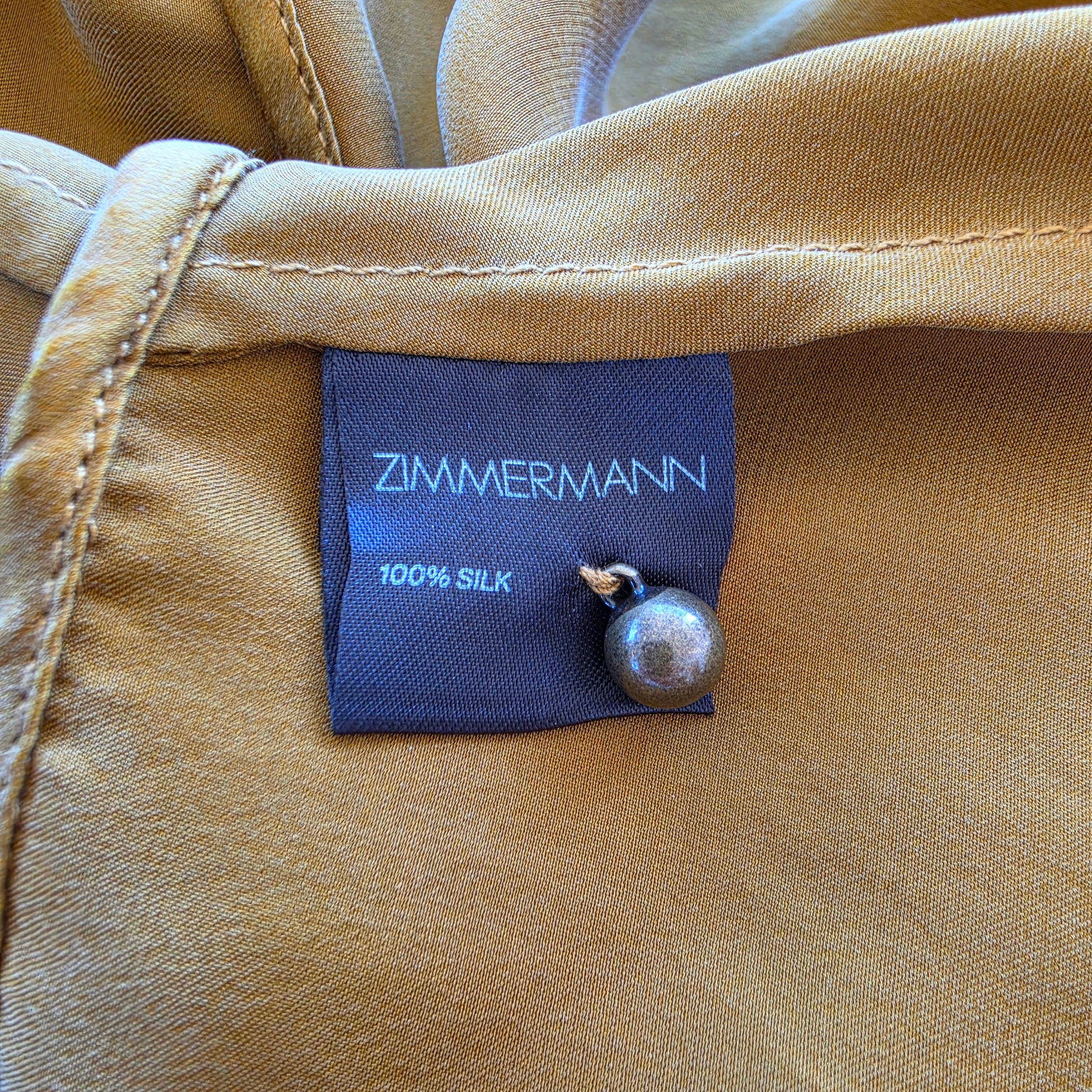 Close-up of the tan garment label reading ZIMMERMANN 100% SILK, with a small metallic pearl pin attached to the side.